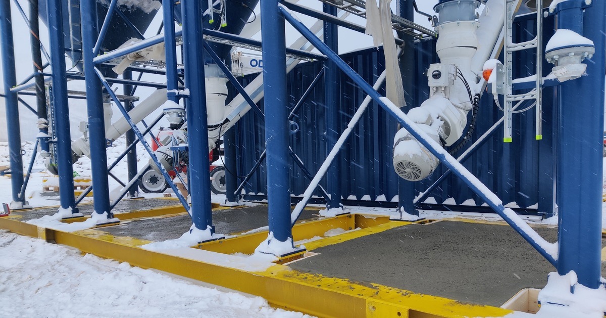 Close-up of a portable concrete plant installed on a mobile steel foundation, showing blue support structures, pipes, and machinery components partially covered with snow.