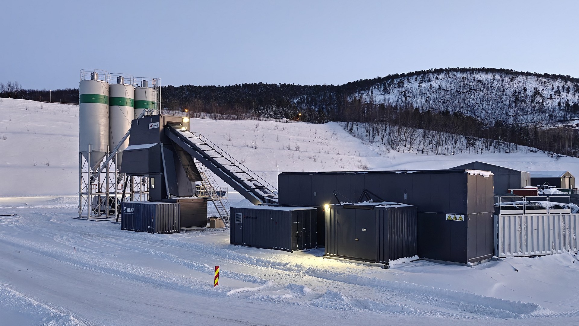 A portable concrete plant installation in a snowy landscape, featuring silos, conveyors, and modular units arranged on a winter construction site with hills in the background.